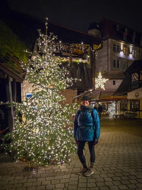 A woman stands outdoors at night near a Christmas tree decorated with white lights. There are holiday-themed lights and decorations, including a star-shaped light, in the background. The setting appears to be a festive holiday scene in a European-sty