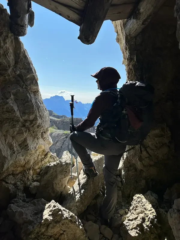 hiker with backpack and trekking poles standing in cave entrance overlooking mountains