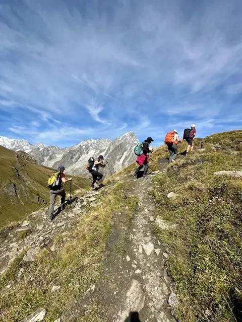 five hikers ascending steep trail with peaks of the alps in the distance while on tour du mont blanc trail