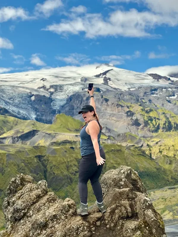 woman standing on rocky point with arms up and snowy mountain in background