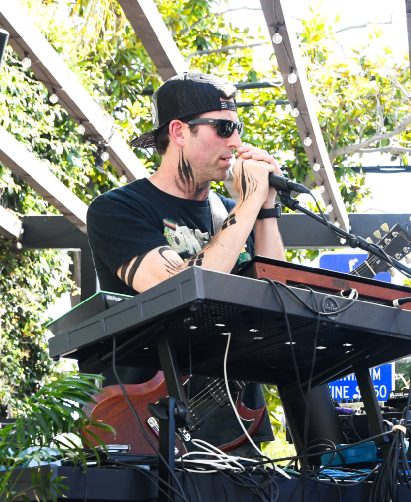 A man is performing music outdoors at a music festival. He is wearing sunglasses, a black cap, and a black t-shirt, with tattoos on his neck and arms. He is singing into a microphone with his hands clasped around it, and there is a keyboard, electronic equipment, and a guitar nearby. The background has greenery and outdoor lighting.
