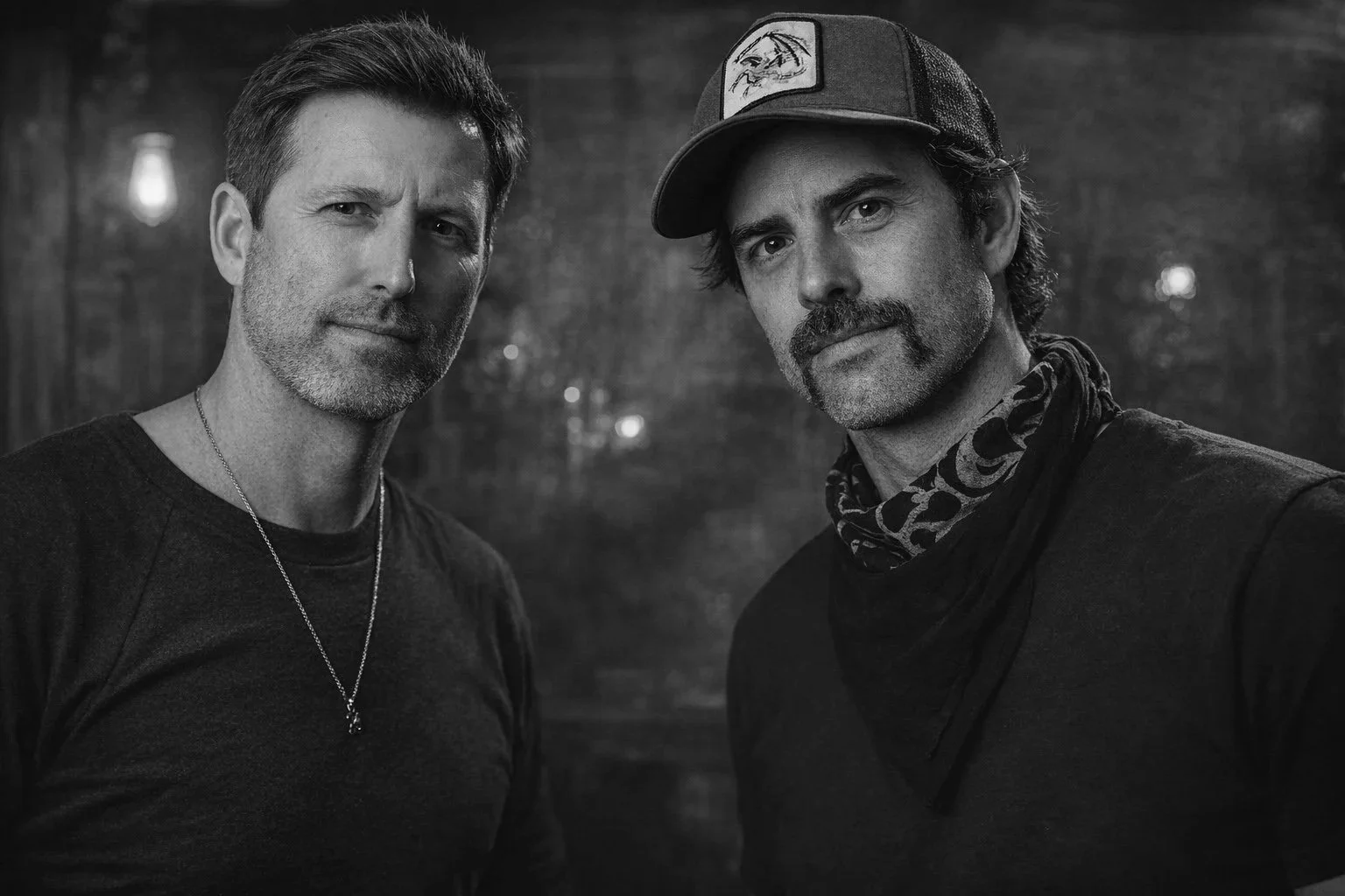 Two men with dark hair and facial hair standing close together outdoors at night, illuminated by soft lighting with trees and hanging lights in the background, in black and white.