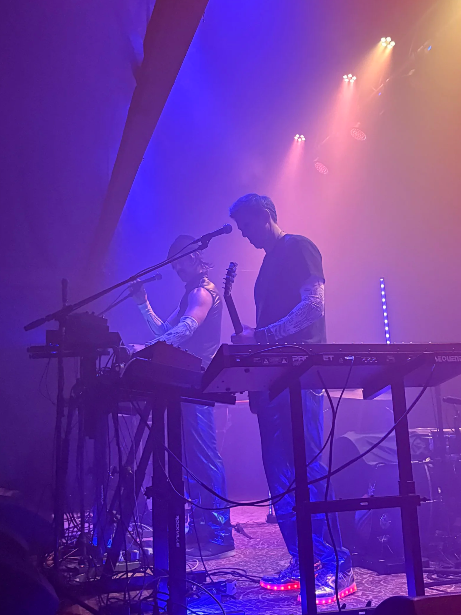 Two musicians performing on stage with colorful purple, pink, and blue lighting, surrounded by musical equipment and cables.