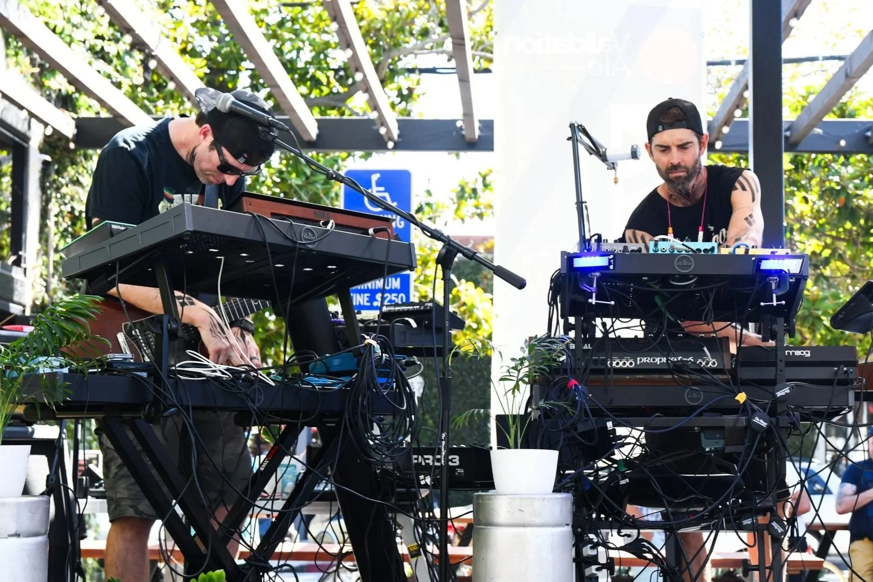 Two musicians performing outdoors with electronic equipment, plants, and trees in the background.