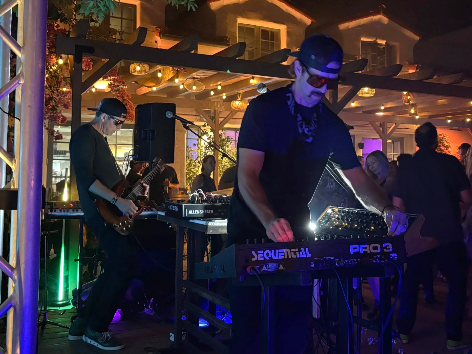 Two musicians performing at a lively outdoor event with colorful lighting, one playing an electric guitar and the other operating a DJ setup, with people dancing in the background.