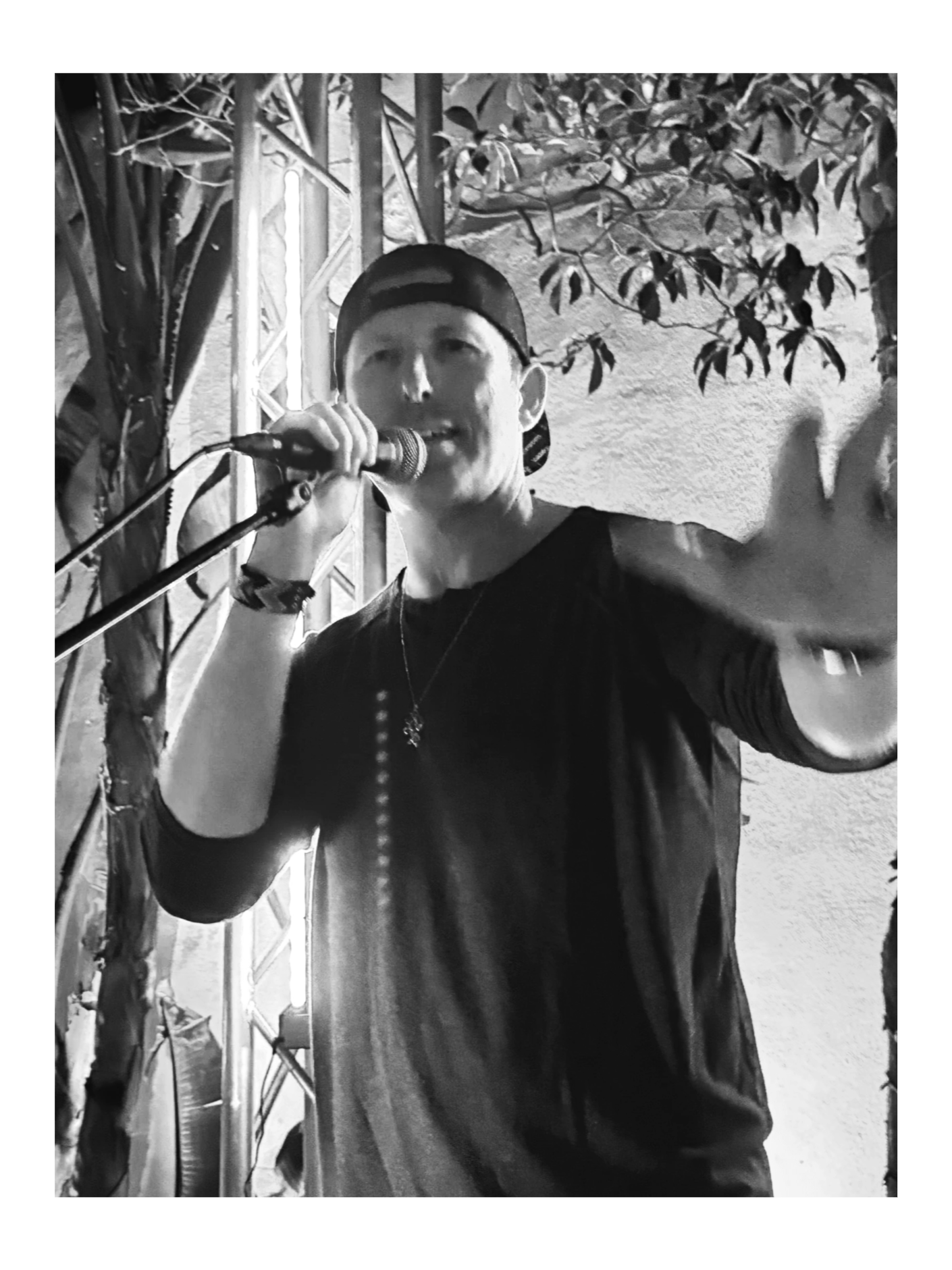 A male performer wearing a backward cap, holding a microphone with his right hand, and gesturing with his left hand, during a performance in a setting with string lights and a plant in the background.