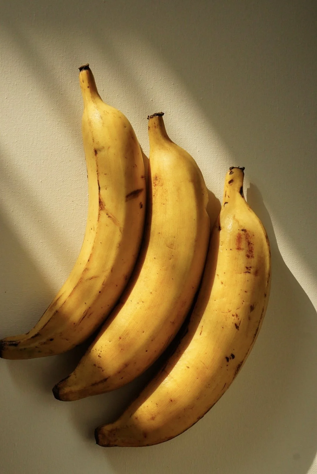 Three ripe bananas on a light-colored surface with a shadow cast on the background.