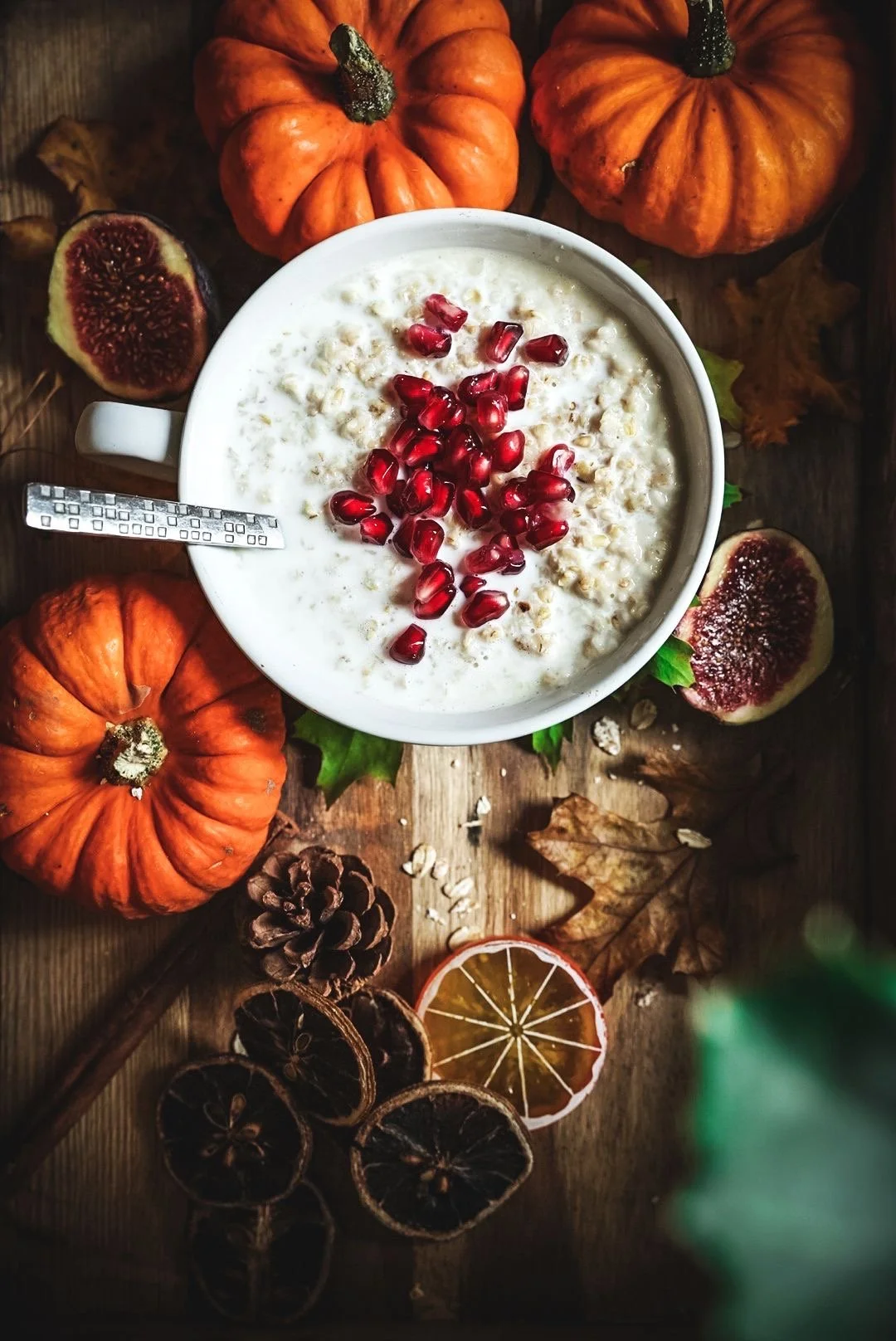 A bowl of oatmeal topped with pomegranate seeds, surrounded by pumpkins, dried figs, dried lemon slices, pinecones, and fallen autumn leaves on a wooden table.