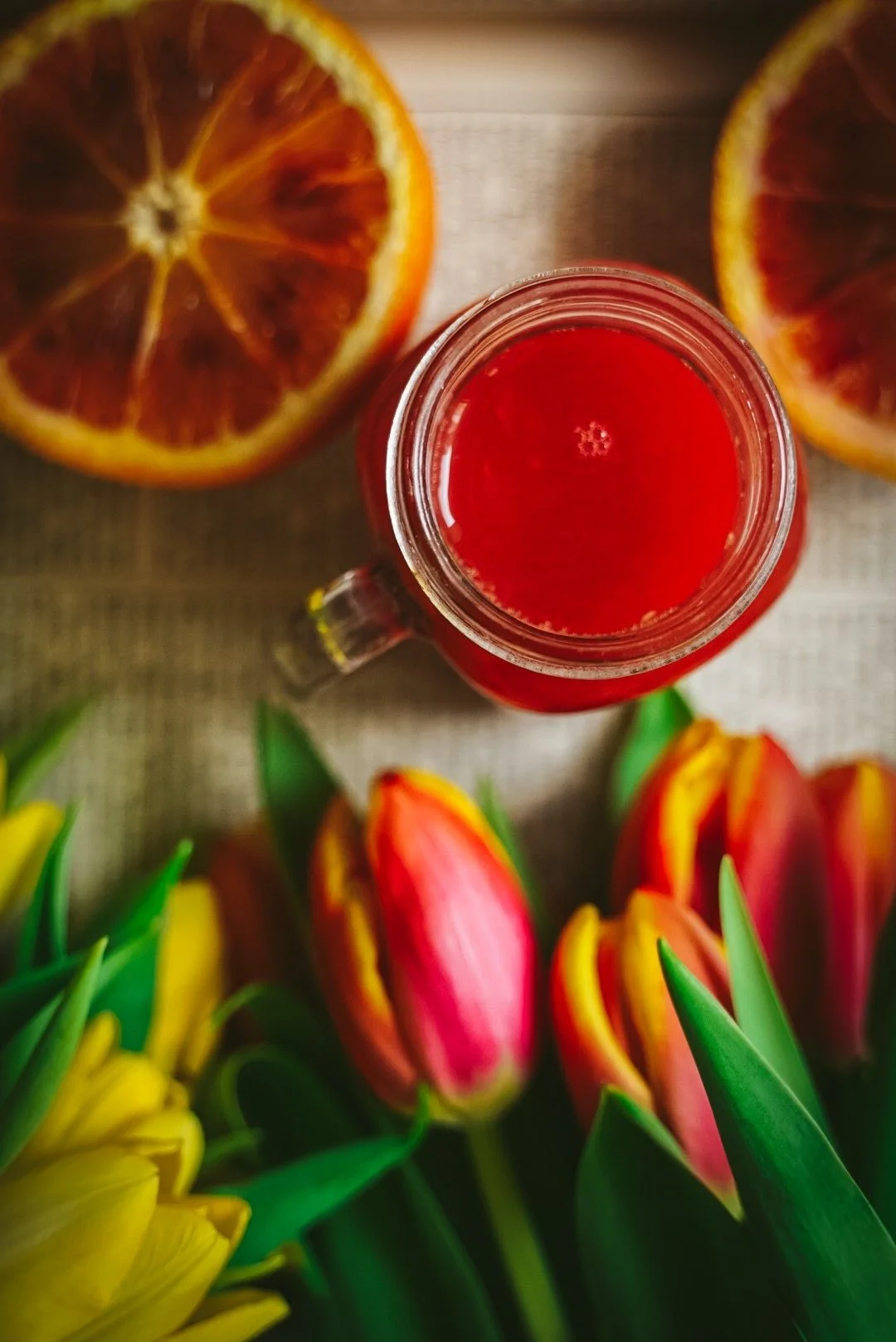 Top-down view of a red beverage in a glass jar with a handle, surrounded by pink and yellow tulips, and half slices of dried blood orange in the background.