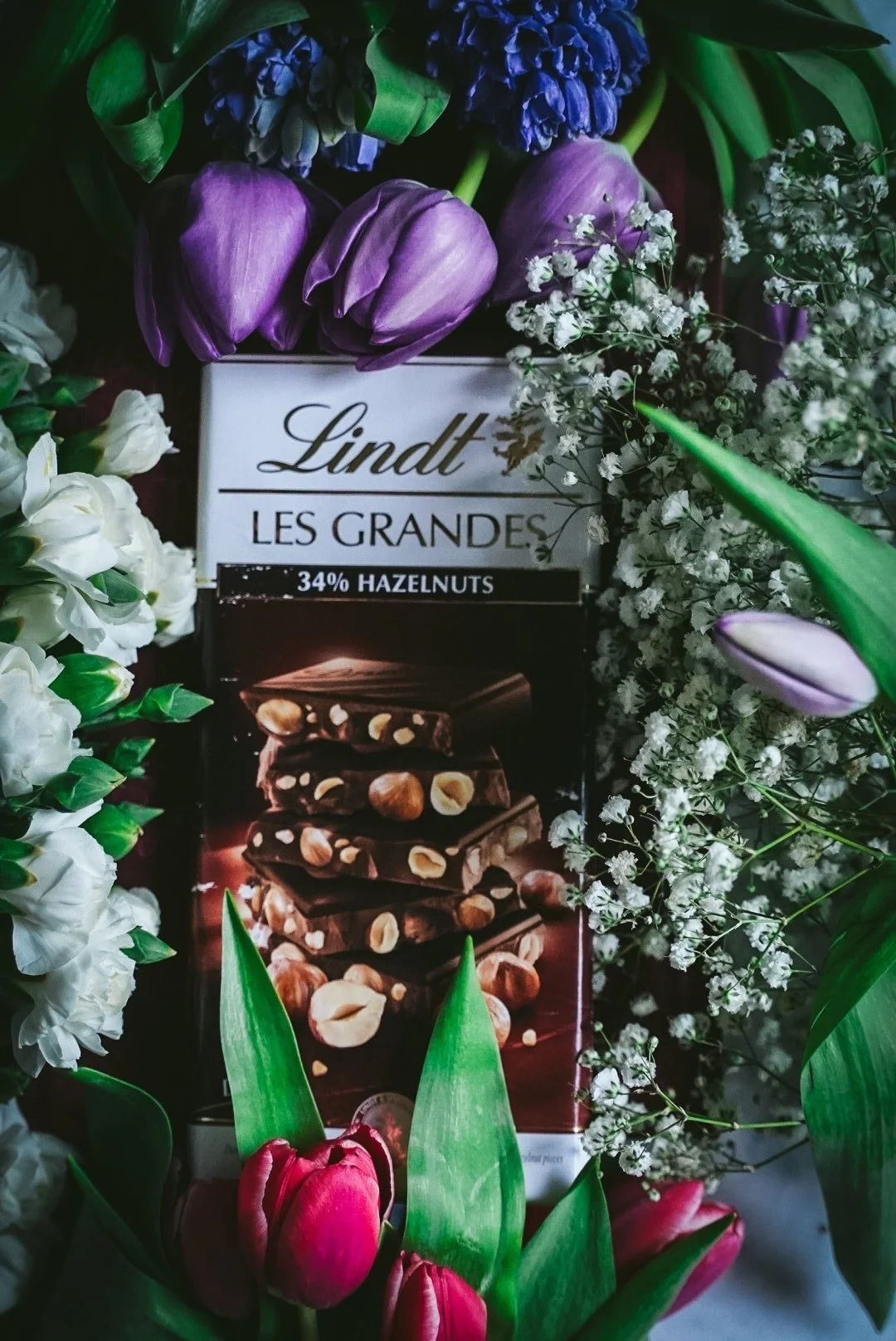 A Lindt Les Grandes dark chocolate bar surrounded by various flowers, including purple tulips, white flowers, pink tulips, and other greenery.