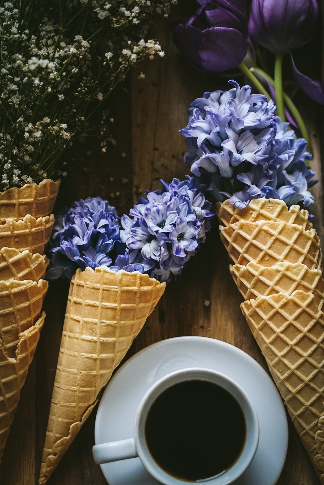 Ice cream cones filled with purple and white hyacinth flowers placed on a wooden surface next to a cup of black coffee and a vase of purple and white flowers.