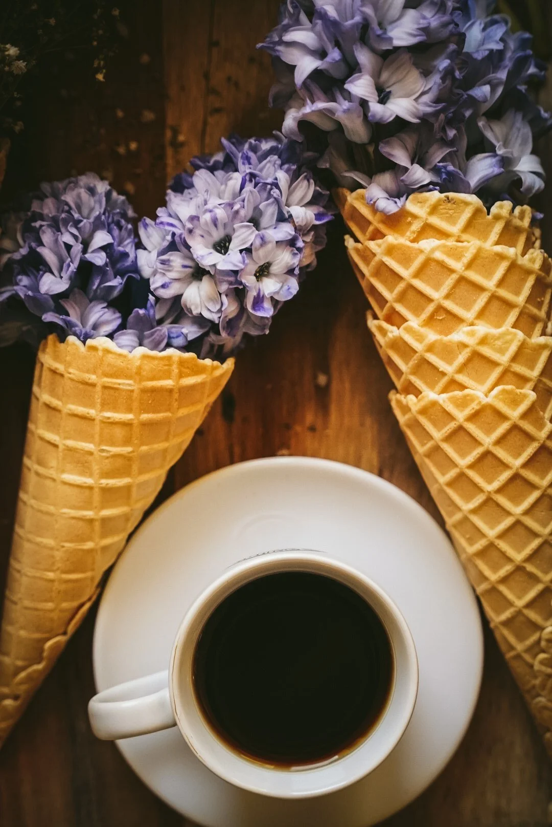 A top-down view of a cup of black coffee on a white saucer, surrounded by three empty waffle cones with purple hydrangea flowers in the cones, set on a wooden surface.
