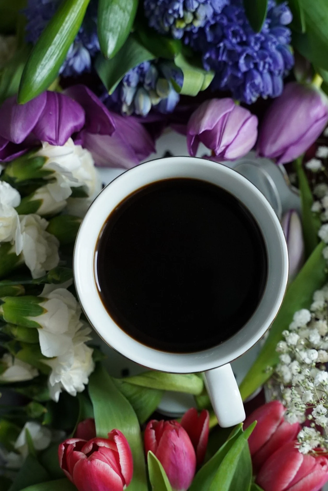 A white coffee mug filled with black coffee surrounded by a variety of colorful flowers, including purple, white, pink, and red tulips, and white baby's breath.