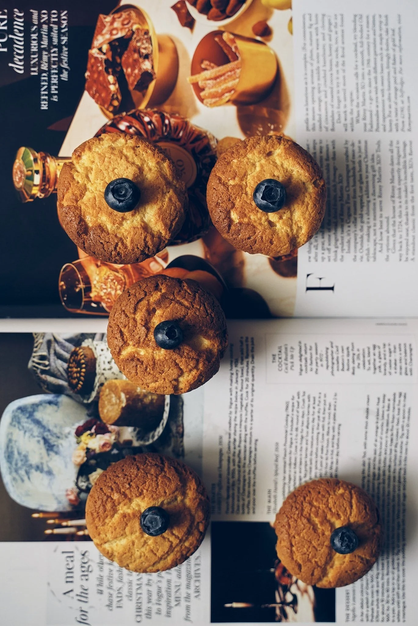 Six muffins topped with blueberries arranged on an open magazine with images of desserts.