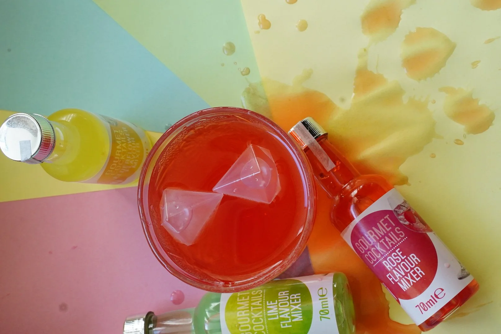 A colorful scene with a pink background, featuring a glass of red liquid with ice cubes, a yellow bottle, a green bottle, and an orange bottle, all labeled as gourmet cocktail mixers with flavors lime and rose.