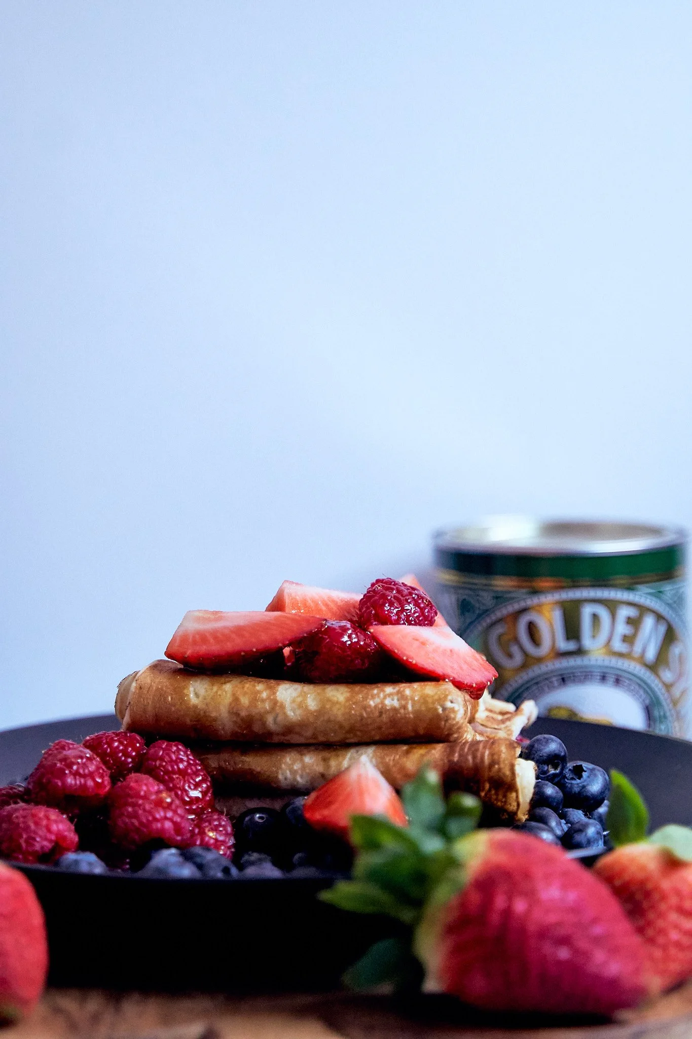 Stacked pancakes topped with strawberries and raspberries, surrounded by additional berries and strawberries, with a can of Golden brand condensed milk in the background.