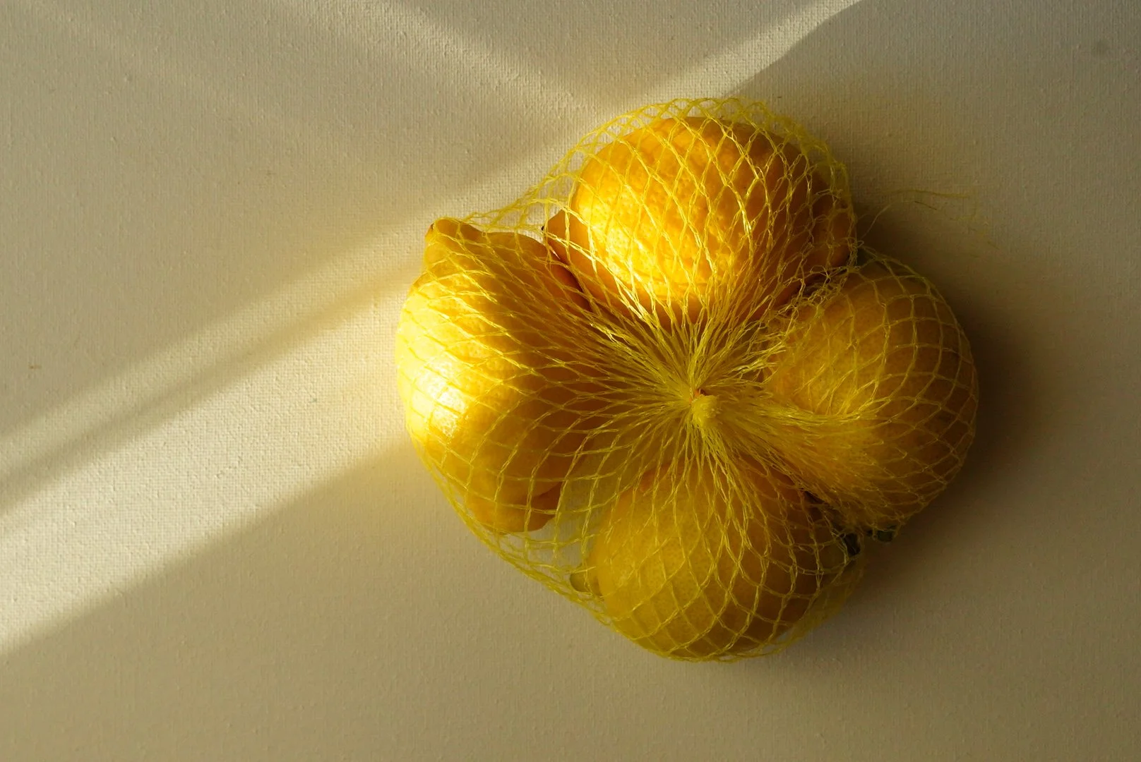 Yellow citrus fruits in a yellow mesh bag, placed on a plain light-colored surface with shadows.