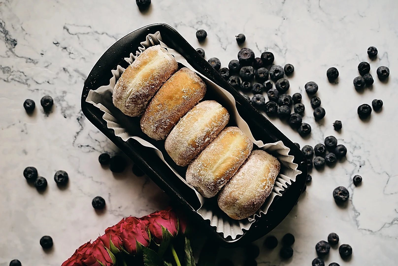 Six powdered sugar-covered pastries in a black container surrounded by scattered blueberries and pink flowers on a white marble surface.