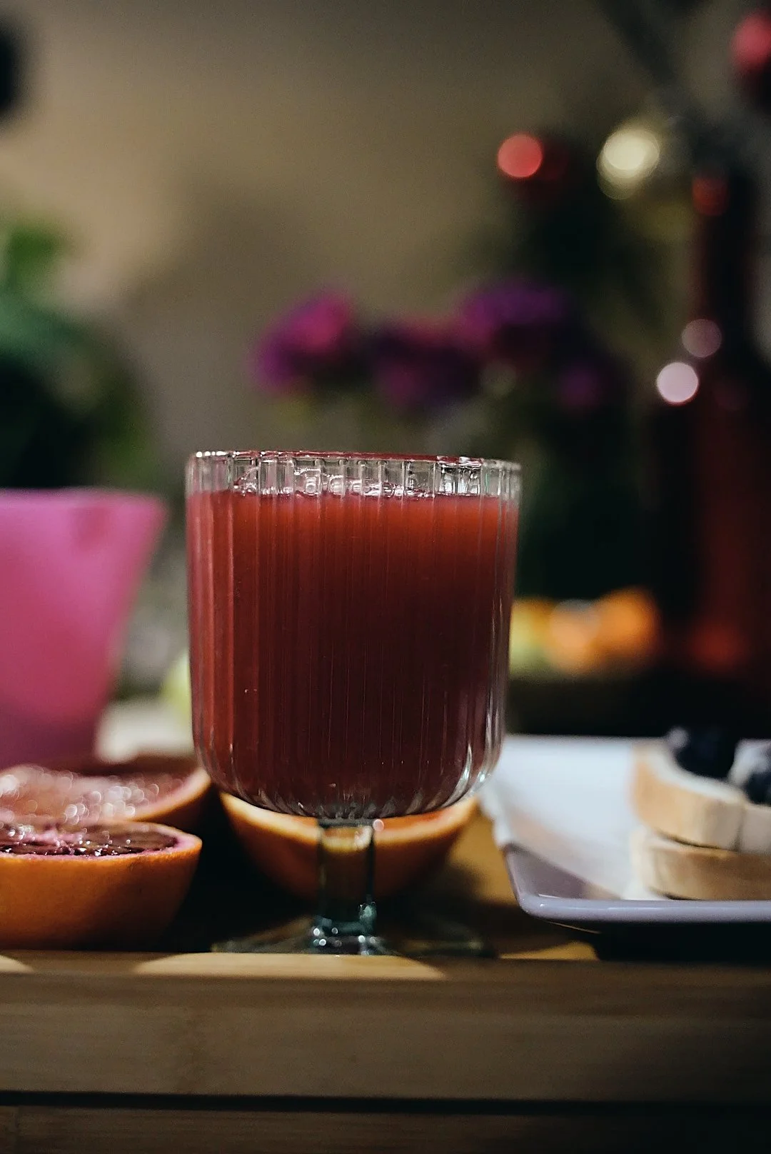 A glass of blood orange juice placed on a wooden table, with halved oranges and a pink flowerpot in the background.