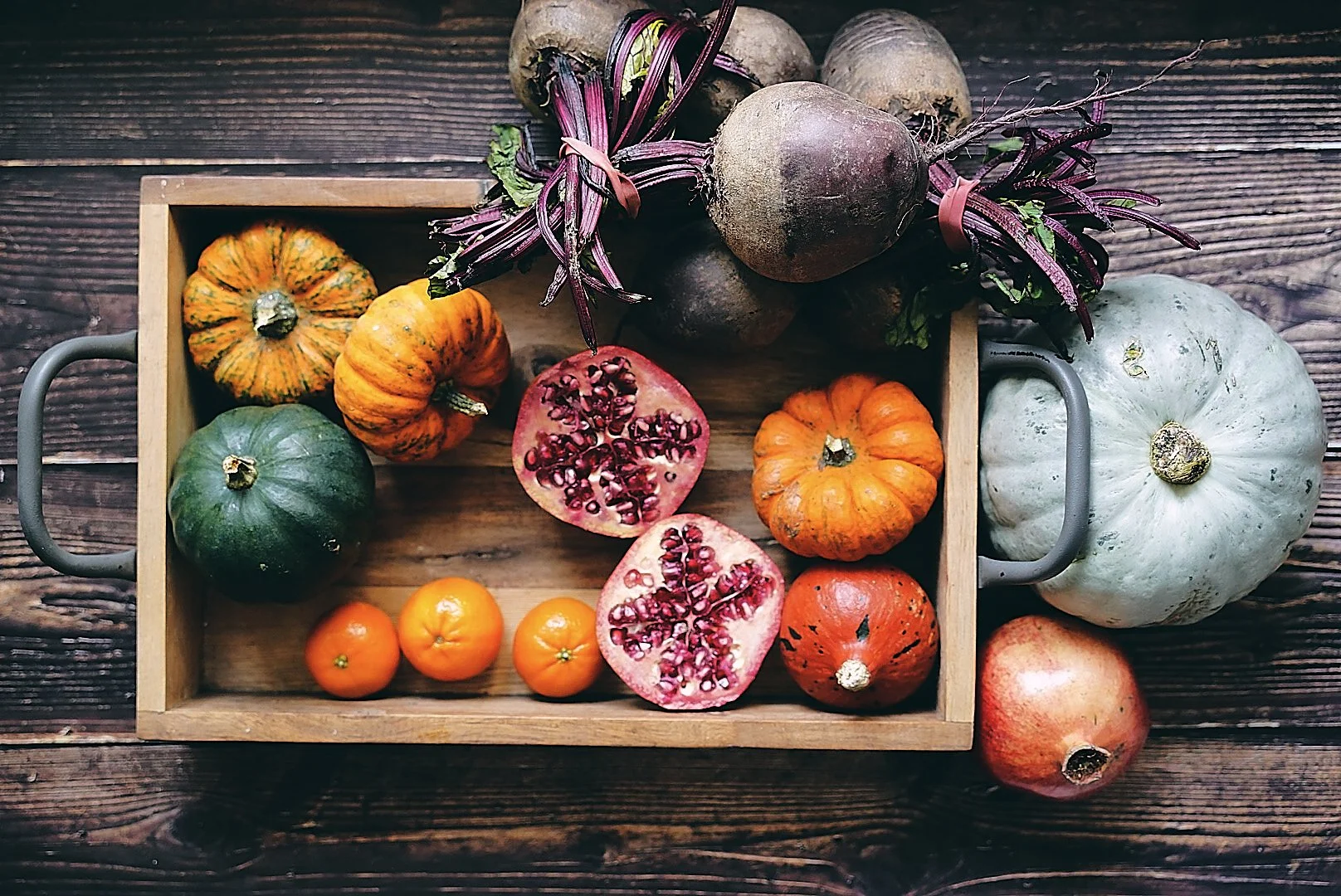 Assorted small pumpkins and gourds in a wooden tray, with a halved pomegranate and purple-tinted leafy branches on a wooden surface.