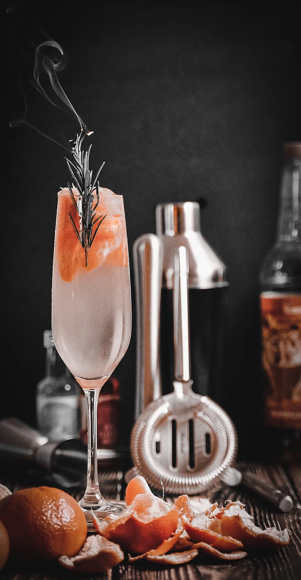 A cocktail in a tall champagne flute garnished with a sprig of rosemary and an orange peel, with a smoky effect on the garnish. The background is dark with bar tools and bottles, and there are peeled oranges and orange peels in the foreground.