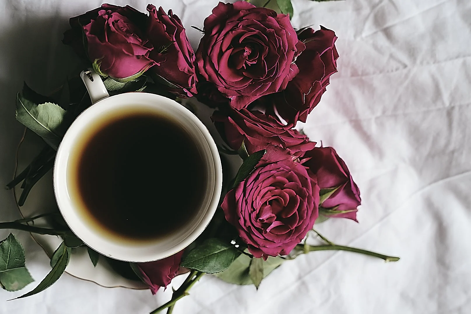 A white mug of black coffee surrounded by pink roses with green leaves, on a white fabric background.