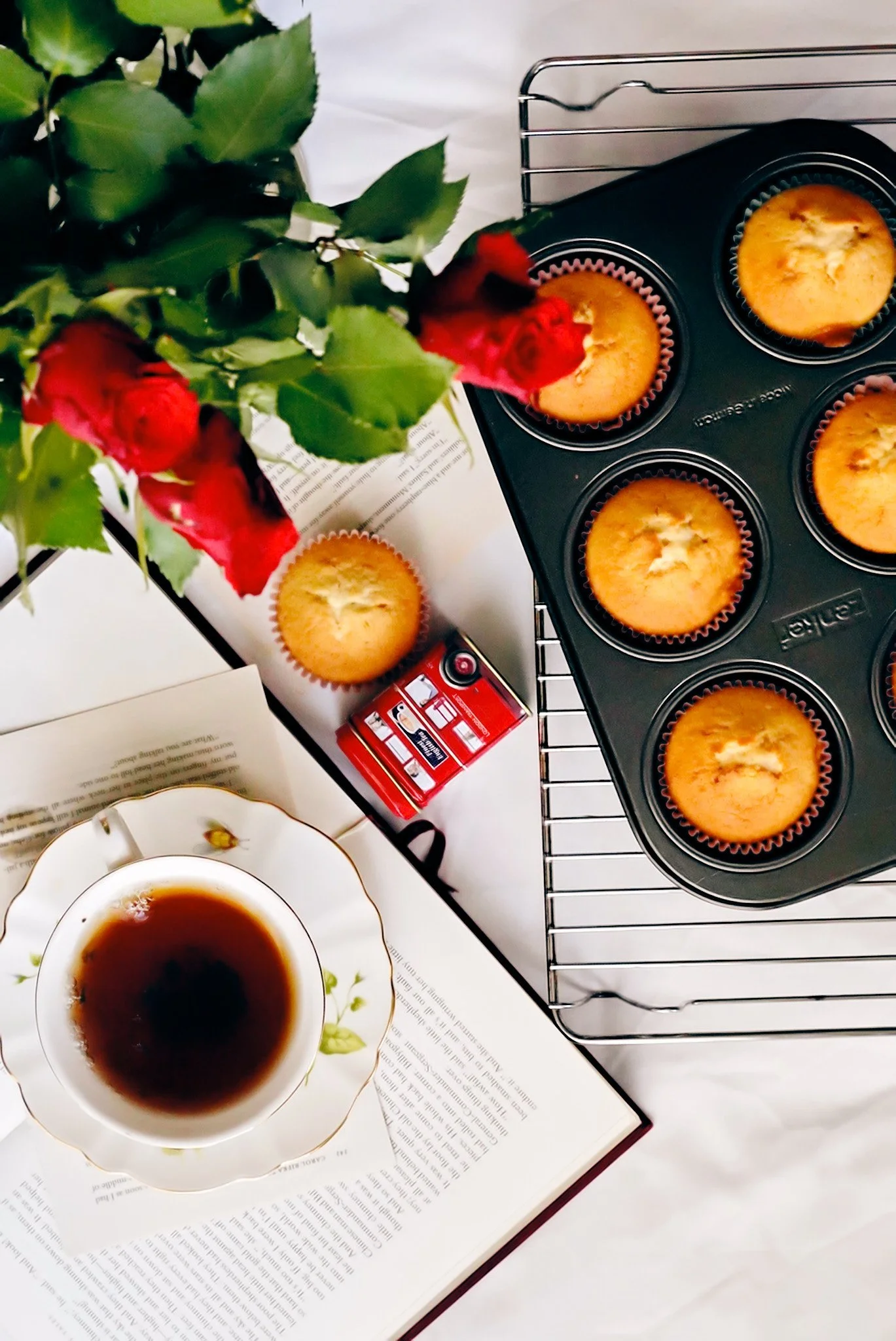 A scene with a cup of coffee on an open book, a tray of six muffins on a cooling rack, a small red toy car, and a red flower in a vase.
