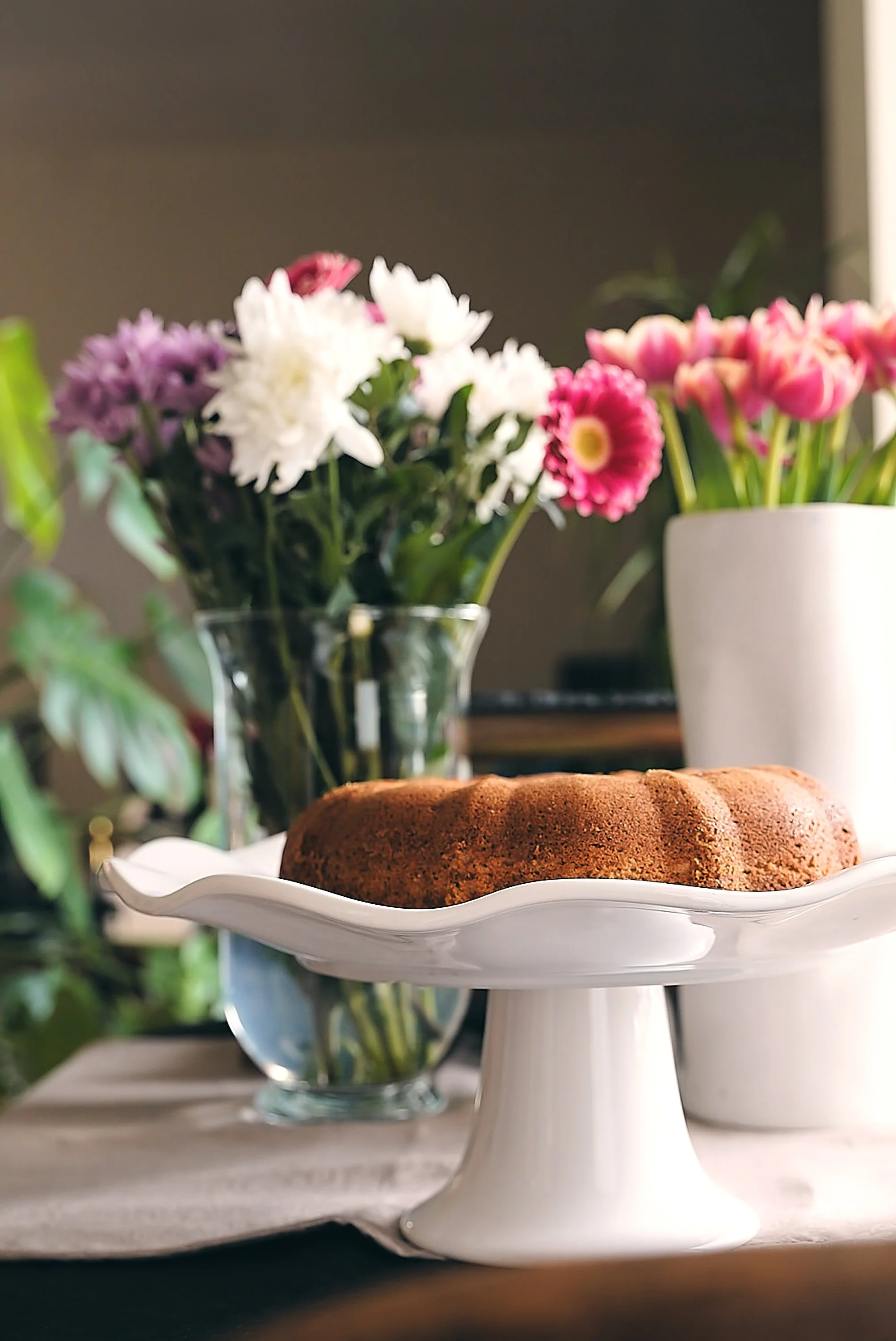 A bundt cake on a white cake stand with flowers in a vase behind it on a table.