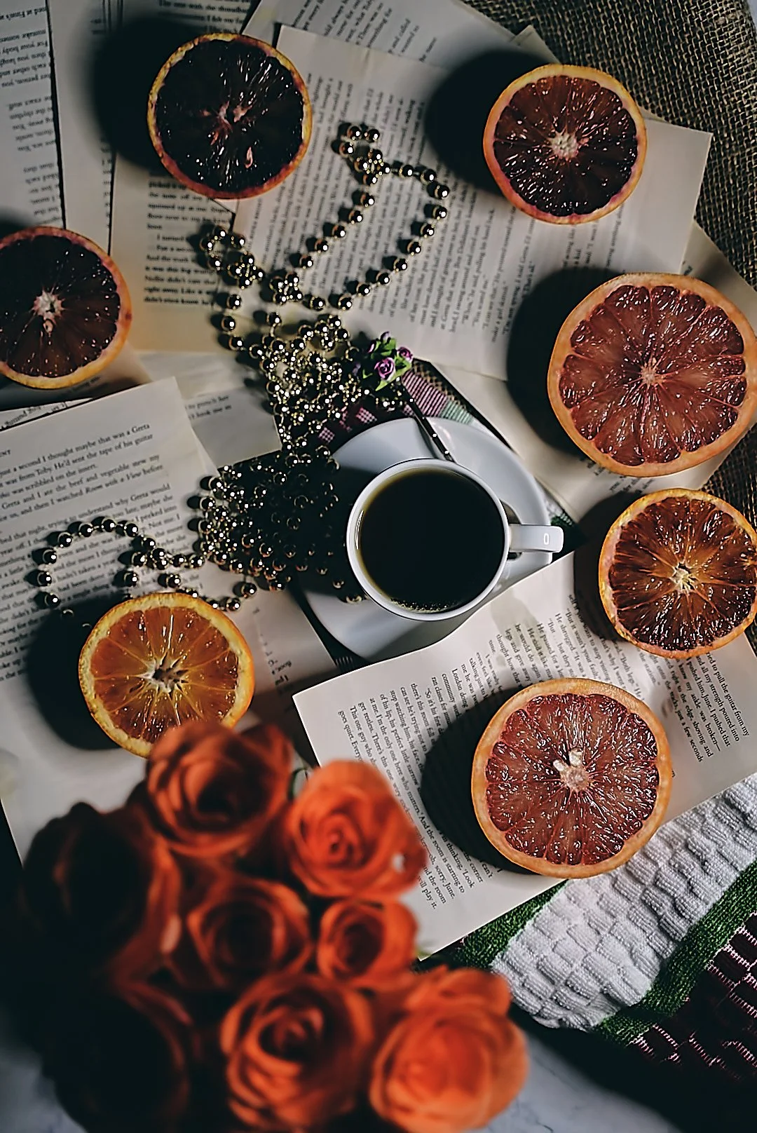 A table scene with halved blood oranges, a cup of black coffee, scattered paragraphs of printed text, black and silver beads, a small purple flower, an orange rose bouquet in the foreground, a white and green cloth, and a manila envelope.