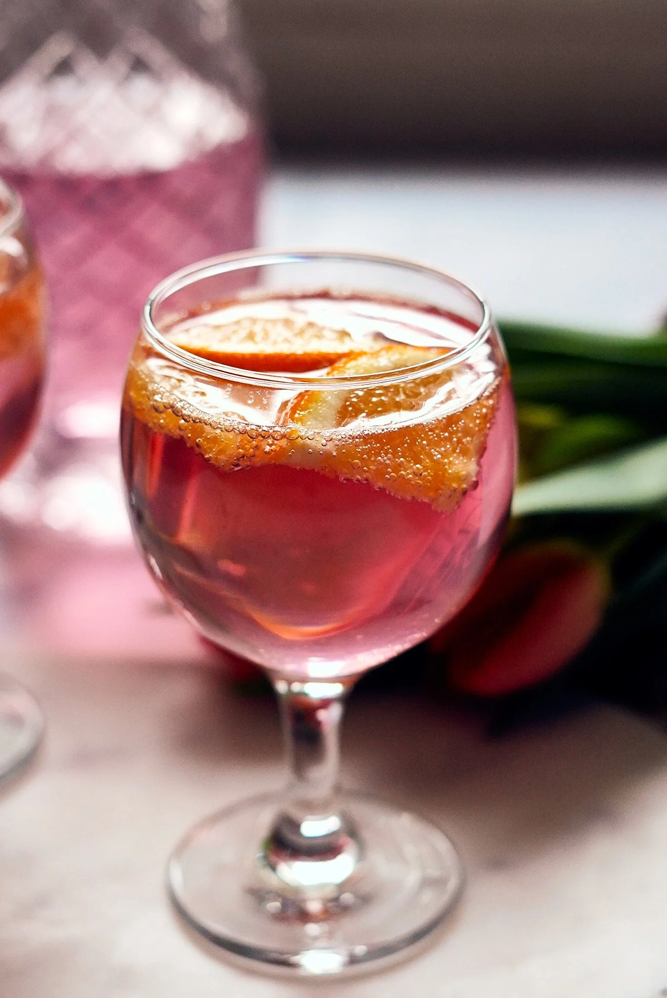 A glass of pinkish cocktail with orange slices. In the background, a pink and purple glass and some green leaves.