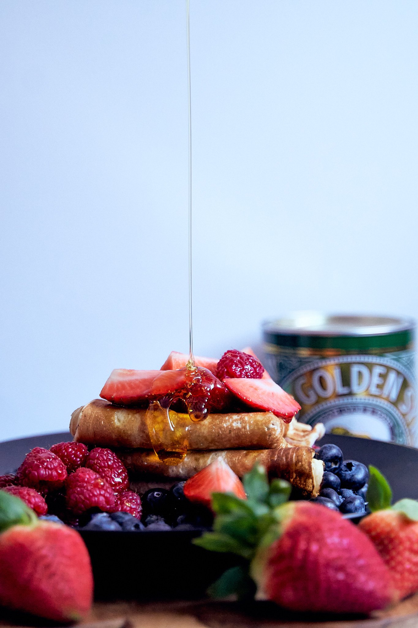Pancakes topped with sliced strawberries and honey, surrounded by fresh berries and strawberries, with an ice cream container labeled "Golden" in the background.
