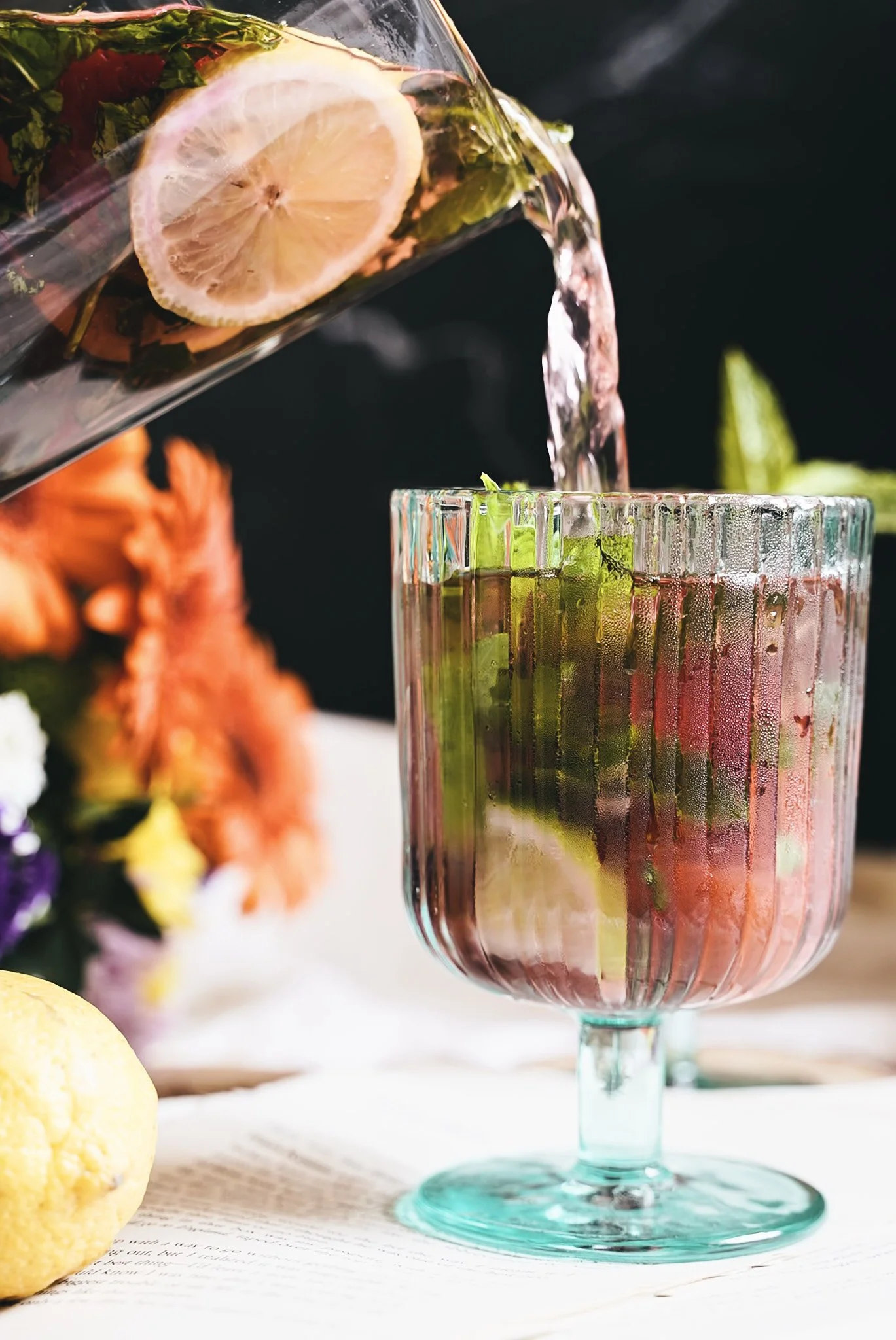 Pouring water with lemon slices and fresh herbs into a glass with ice and lime slices on a table with flowers and a lemon