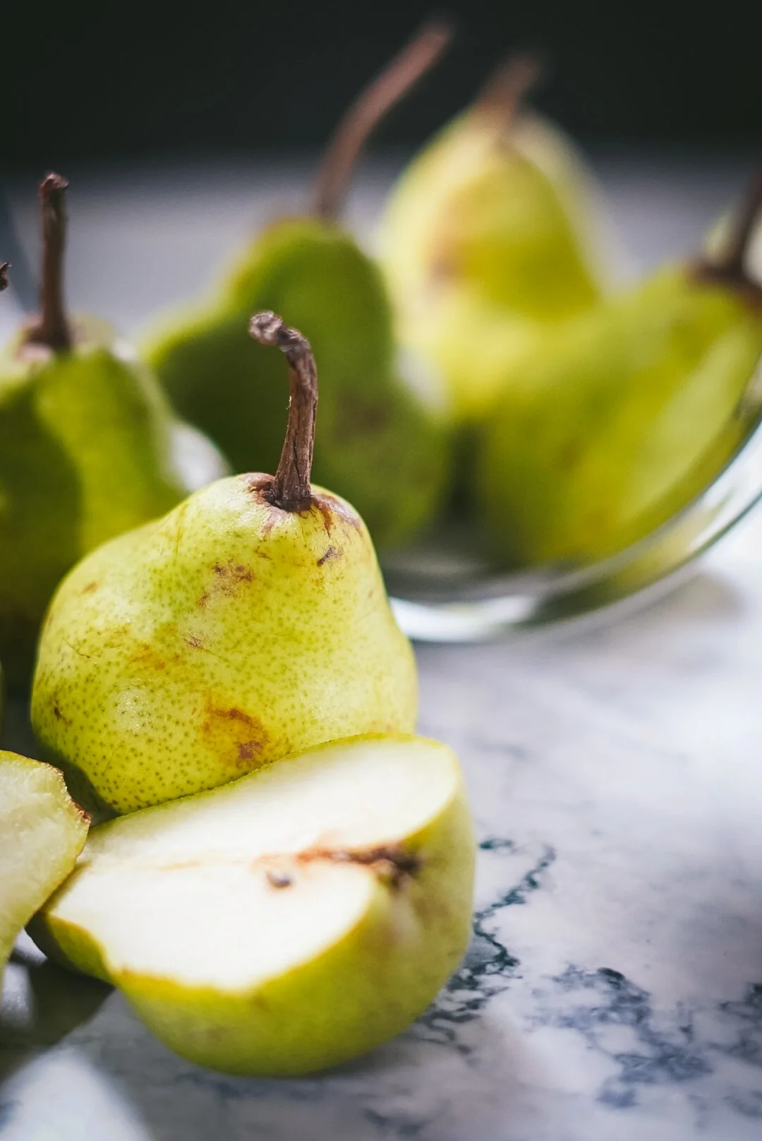 Fresh green pears with one sliced open on a marble surface, with a bowl of more pears in the background.