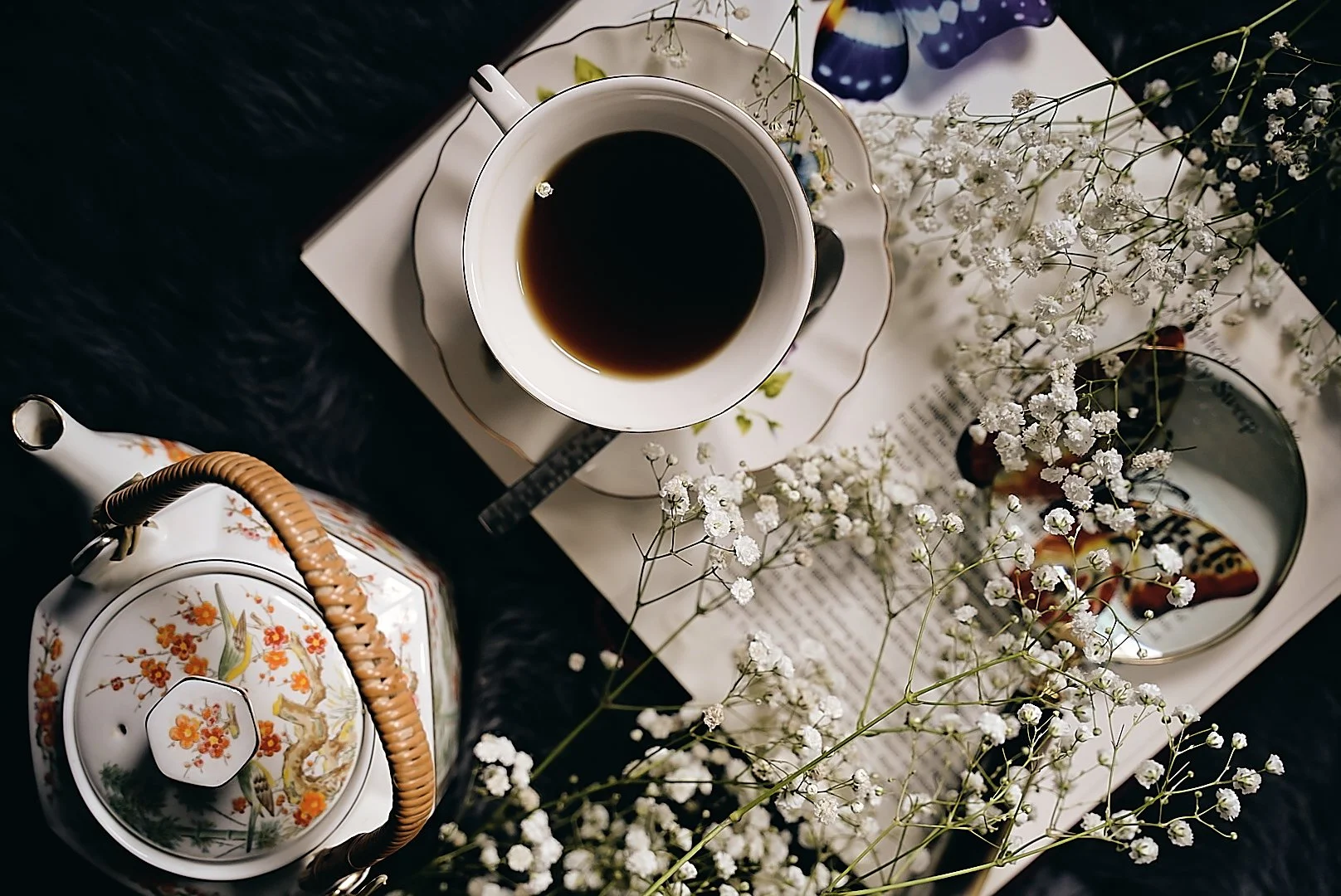 A top-down view of a tea setting with a cup of black coffee on a saucer, a teapot decorated with flowers, white baby's breath flowers, a mirror with butterfly-shaped objects, a magazine or newspaper, and a black surface underneath.