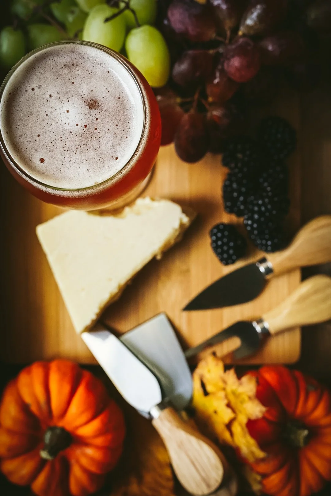 A glass of beer with foam on top, surrounded by grapes, blackberries, a slice of cheese, pumpkins, and cheese graters on a wooden surface.