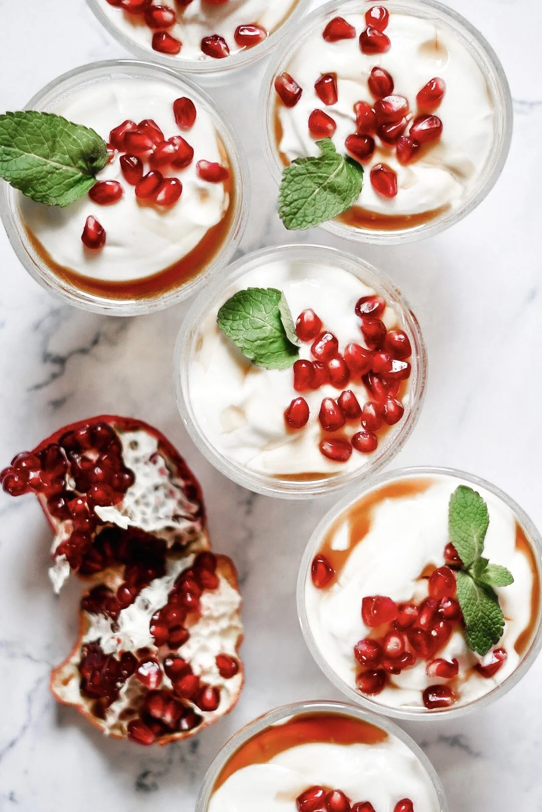Six cups of dessert topped with pomegranate seeds and mint leaves, with a partially eaten pomegranate on the side.