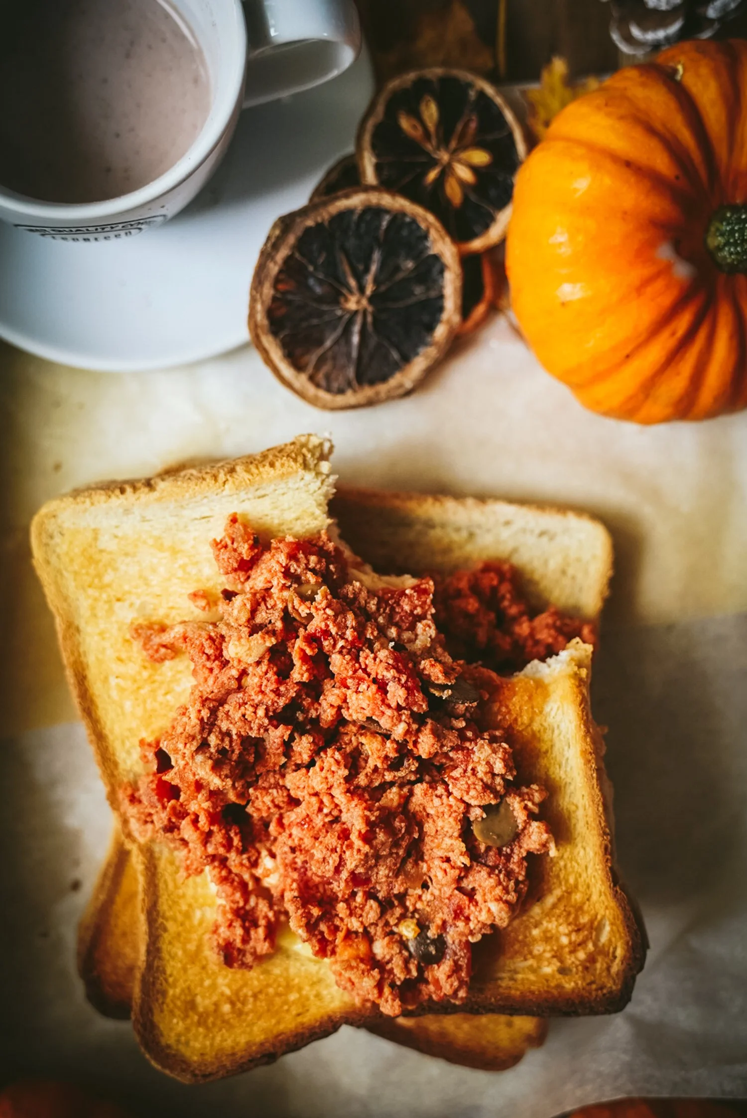 A breakfast scene featuring a toasted bread with a meat spread, a cup of coffee, dried lemon slices, a small pumpkin, and a small glass of syrup or honey.