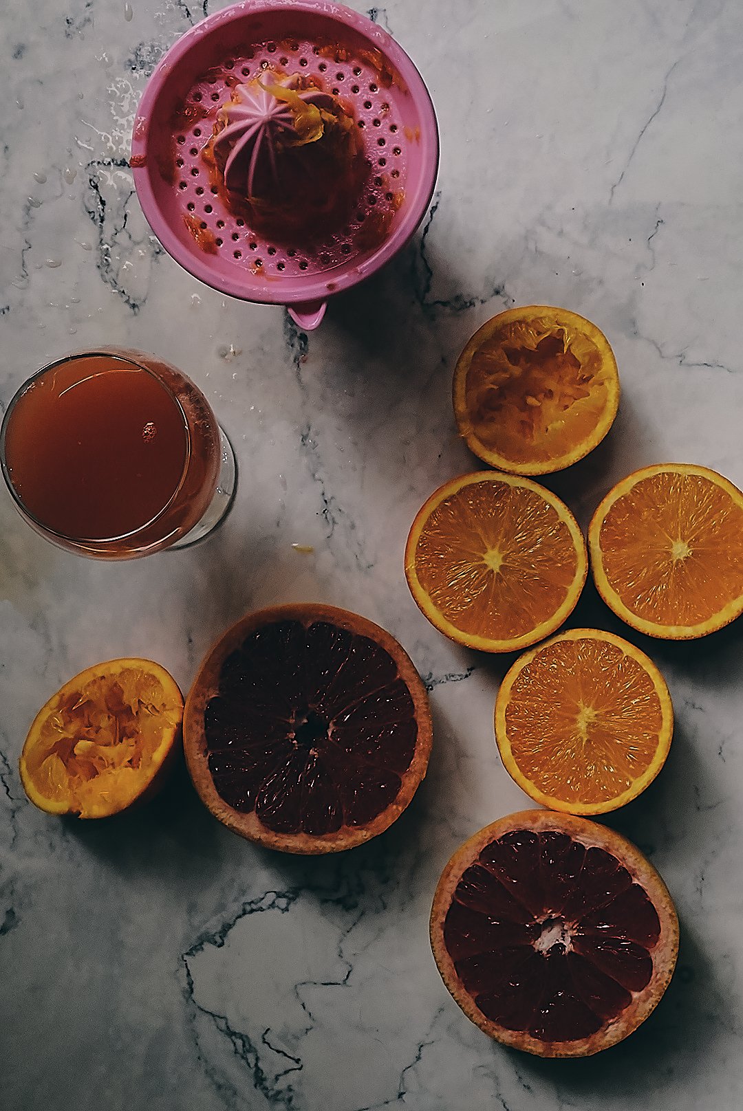 A marble surface holding halved citrus fruits, including oranges, grapefruits, and lemons, with a pink plastic cup and a glass of tea.