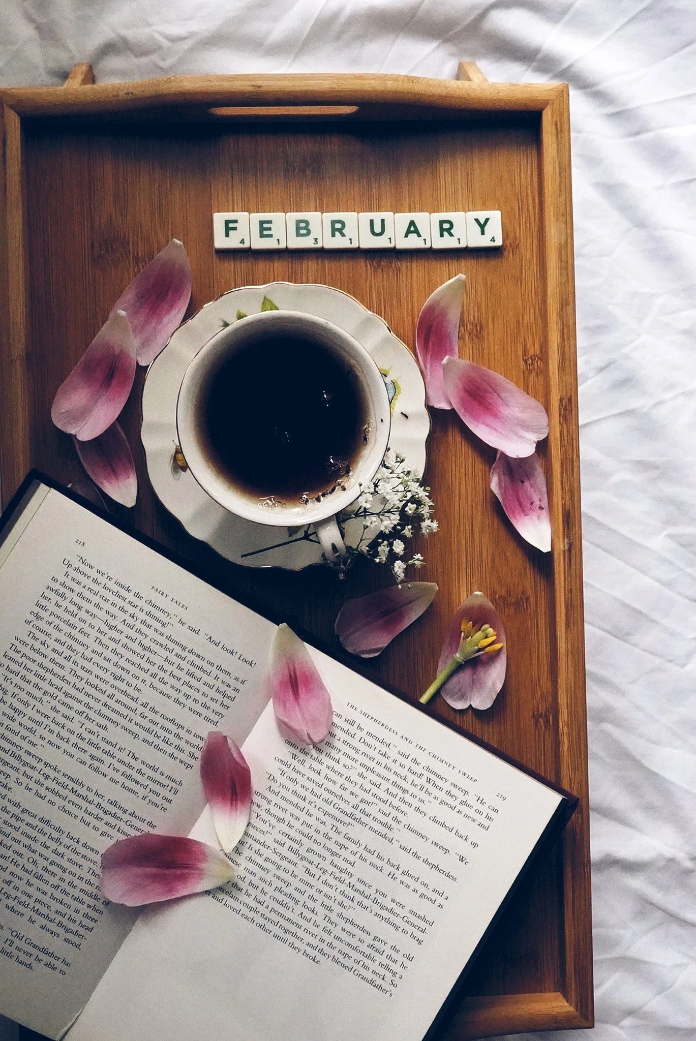 A wooden tray holding a cup of coffee, pink flower petals, an open book, a small flower, and Scrabble tiles spelling "February."