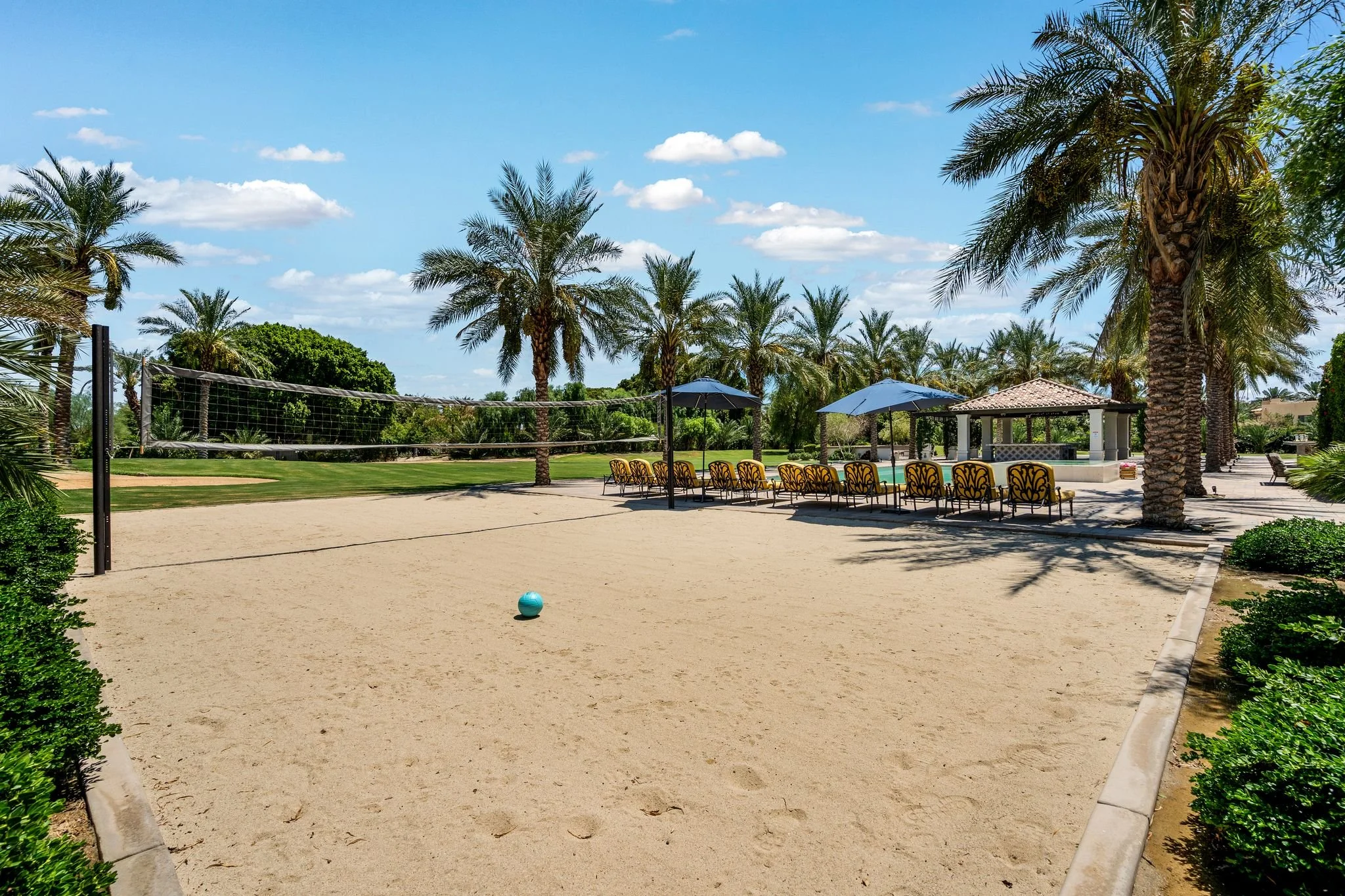 A sandy beach volleyball court with a volleyball, surrounded by palm trees, lounge chairs, and umbrellas under a blue sky with scattered clouds.