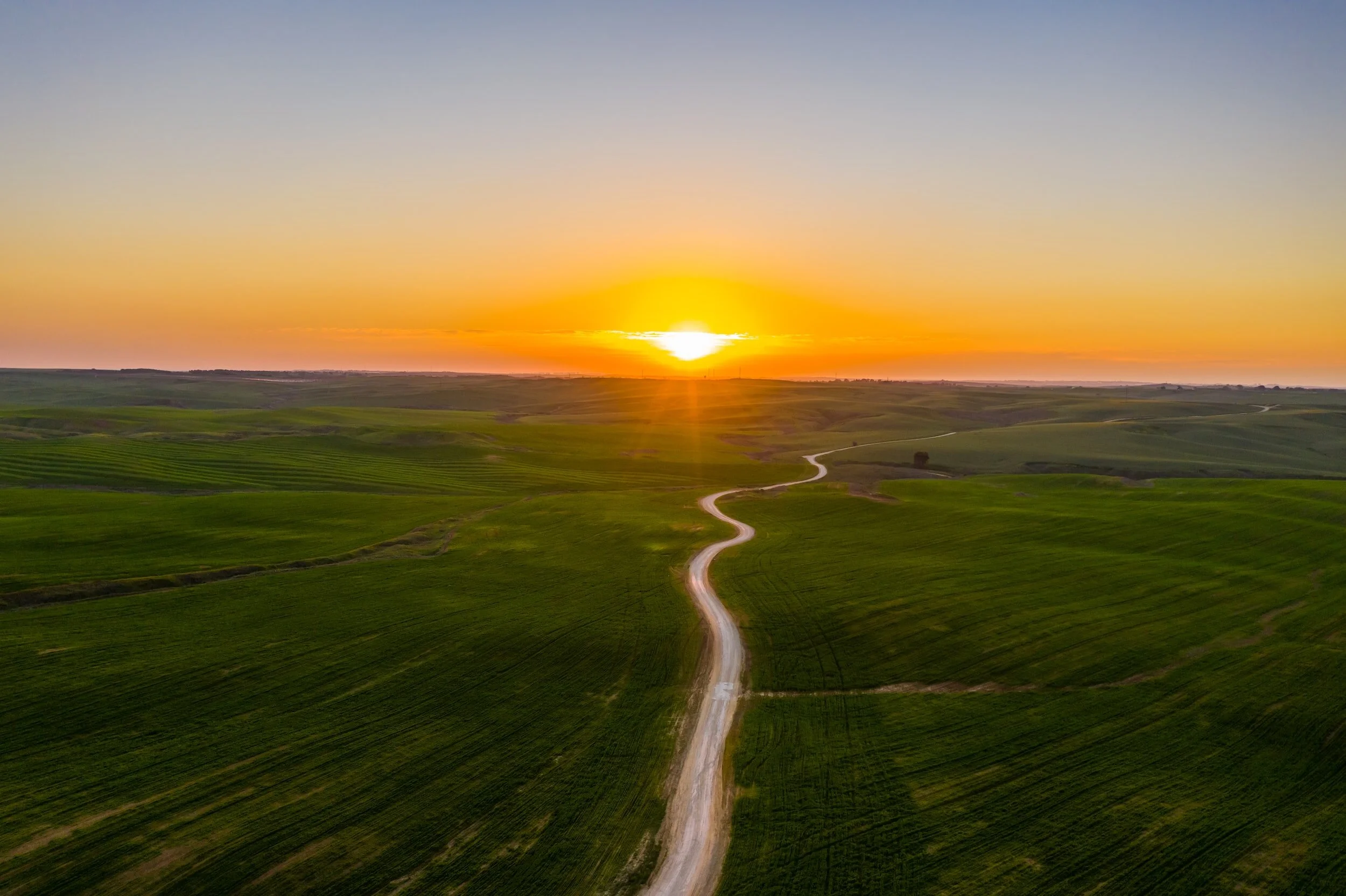 Landschaft mit einer gewundenen Straße, grünen Feldern und Sonnenuntergang am Himmel.