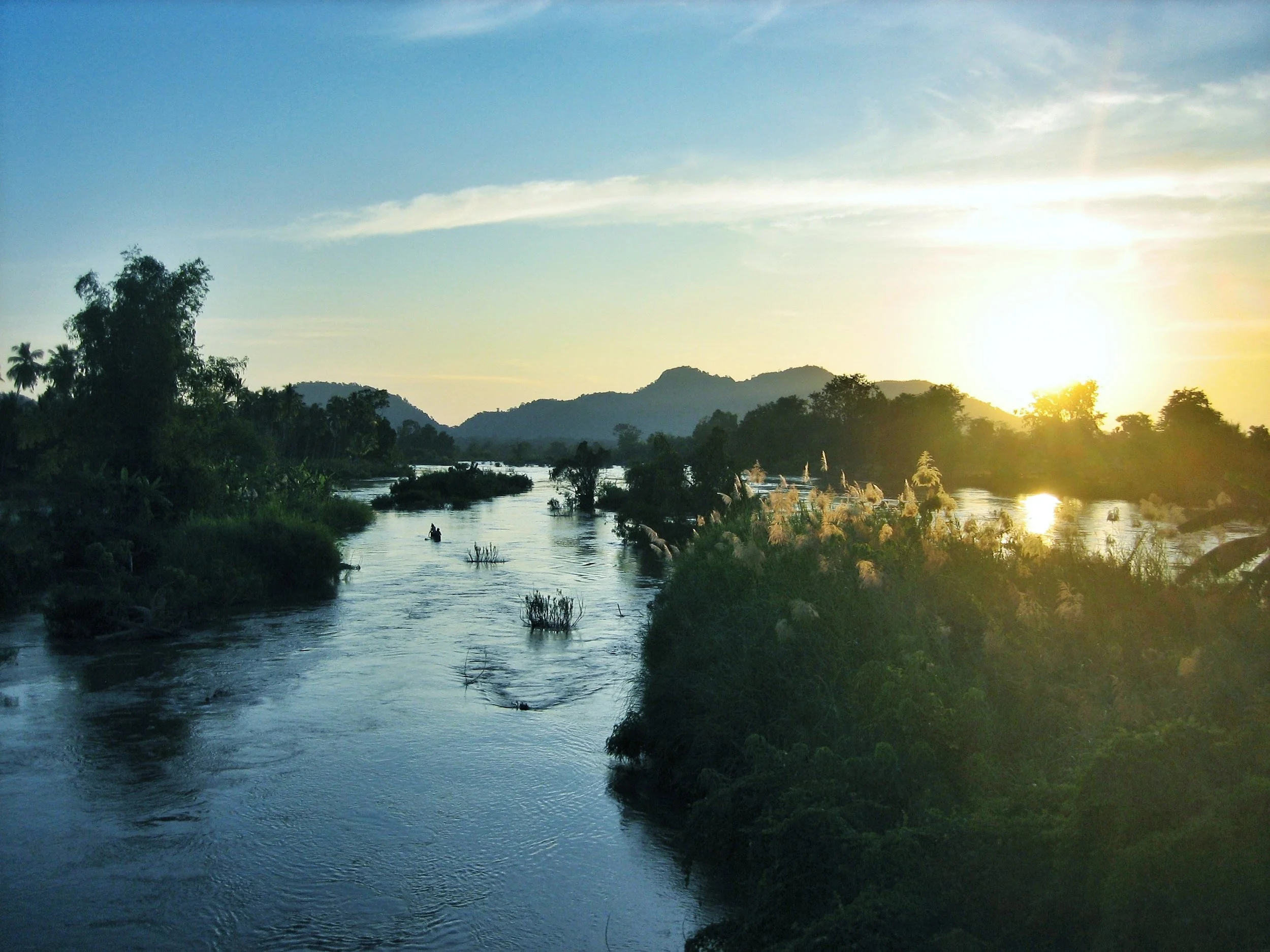 Ein Fluss fließt durch eine grüne Landschaft bei Sonnenuntergang, mit Hügeln im Hintergrund und Bäumen entlang des Ufers.