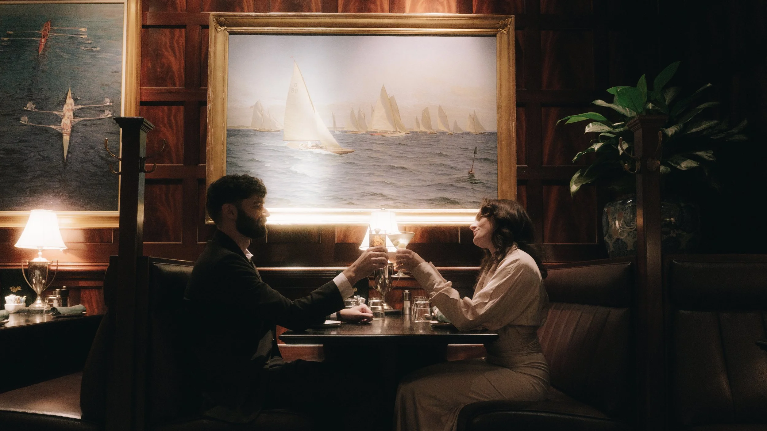 A couple sitting at a dinner table in a dimly lit restaurant, clinking glasses, with large maritime paintings on the wooden-paneled wall behind them.