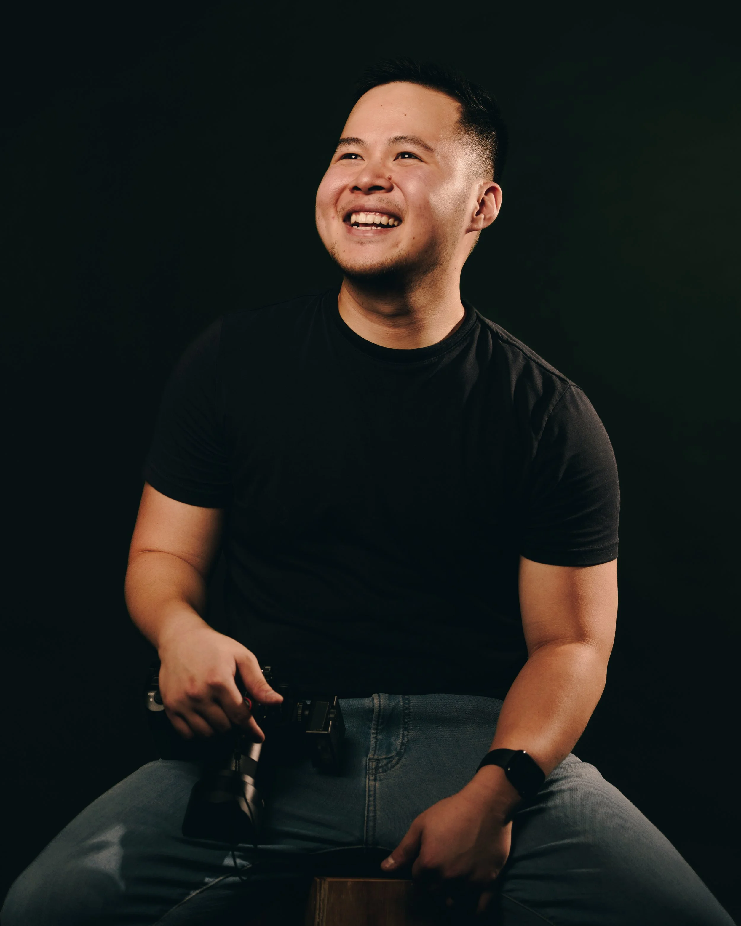 A young man smiling and looking upwards, holding a camera, wearing a black t-shirt and jeans, seated against a dark background.