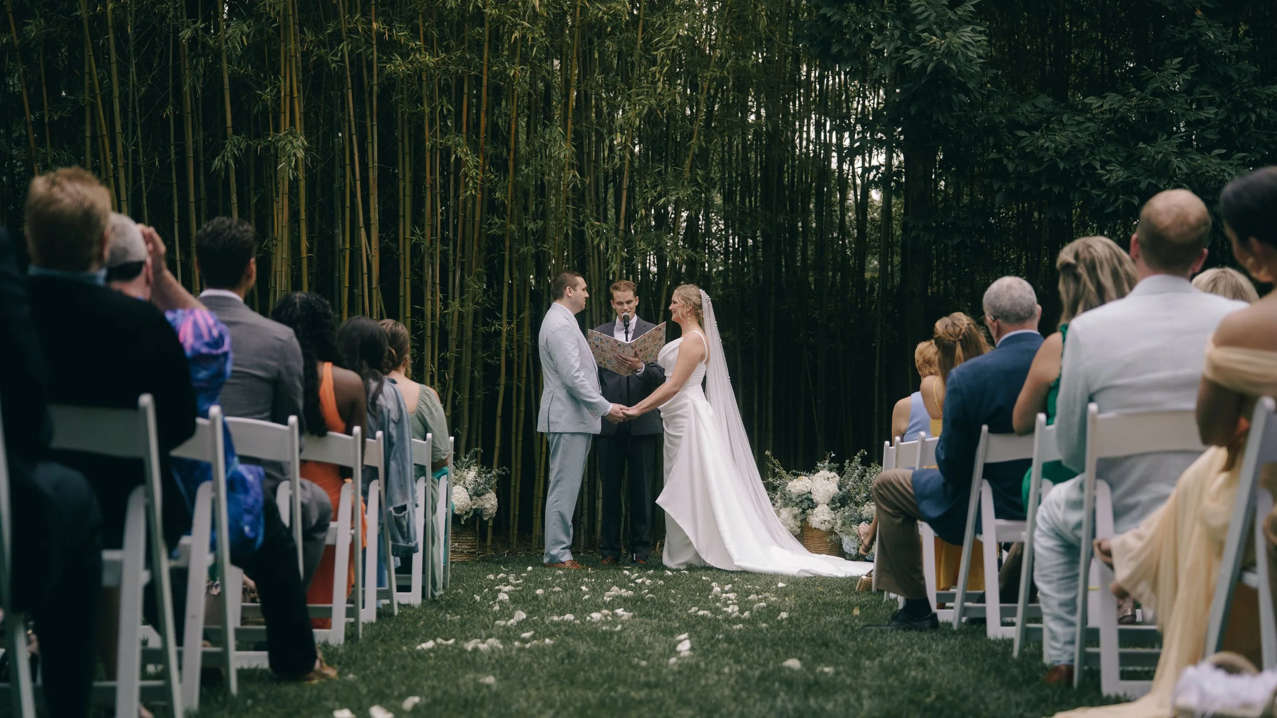 A couple gets married outdoors in a garden with bamboo trees in the background, holding hands and facing each other in front of an officiant, surrounded by seated guests.