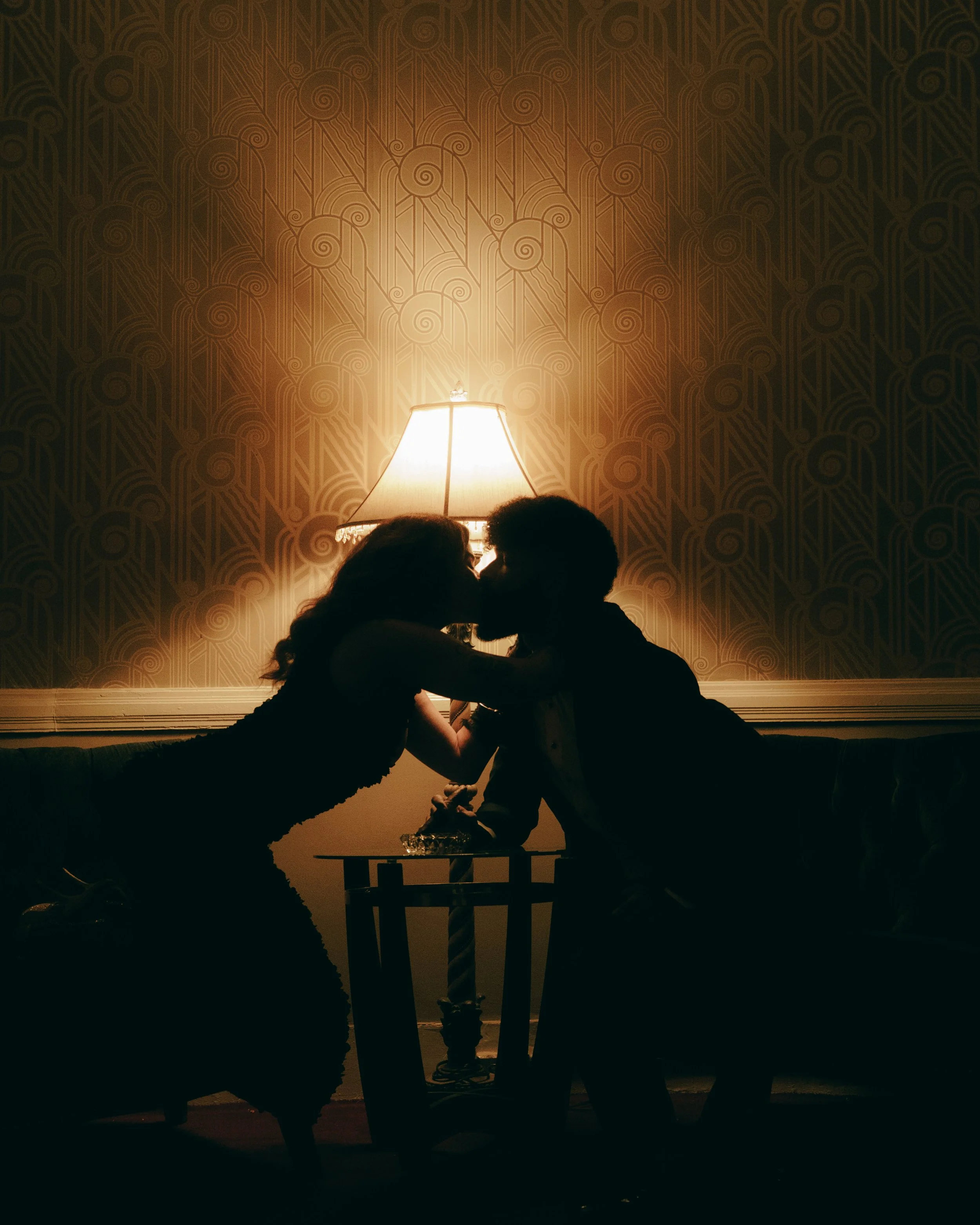 Silhouette of a couple kissing in a dimly lit room with a table and a lamp behind them.