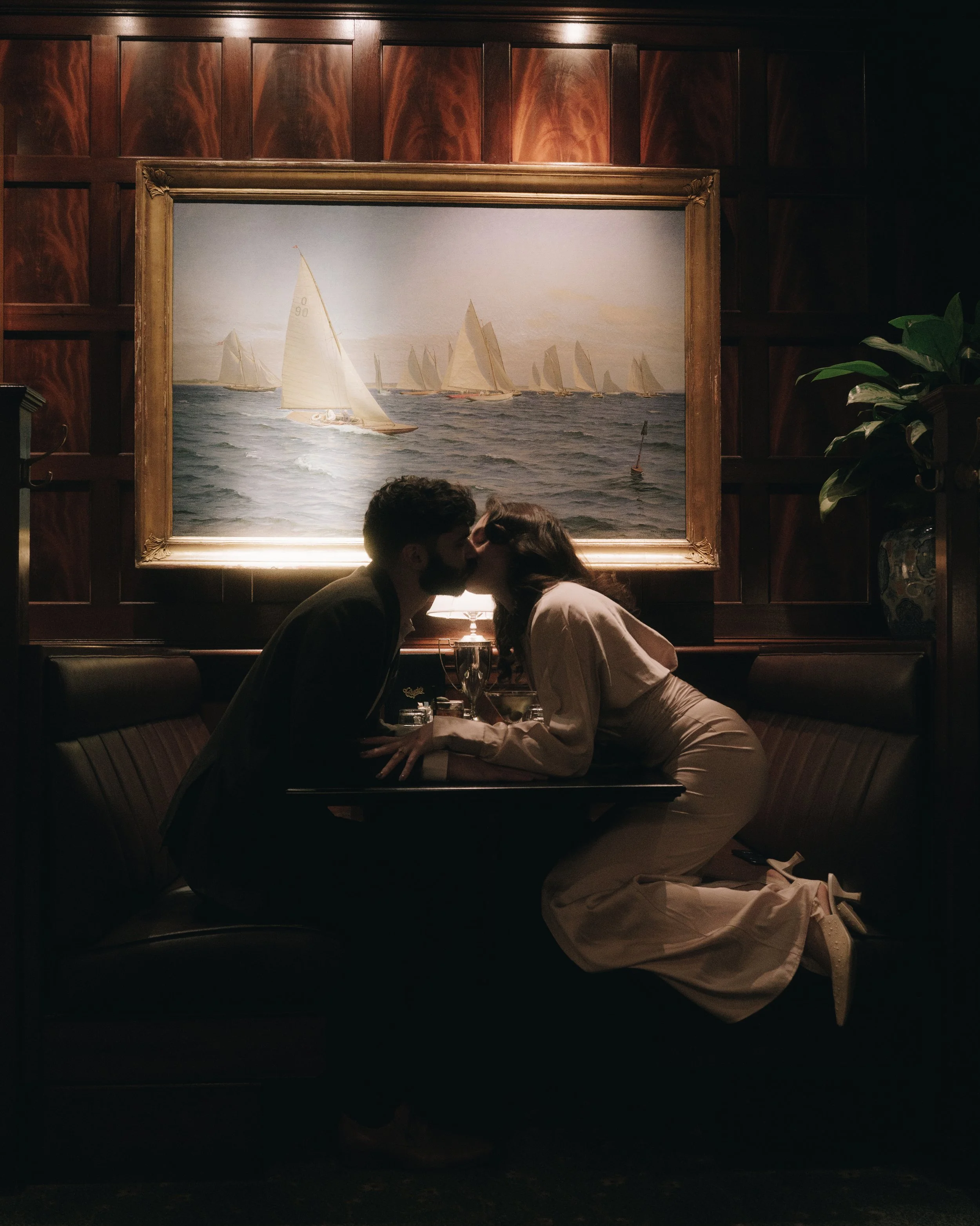 A couple sharing a kiss at a restaurant table with a painting of sailboats on the ocean hanging behind them.
