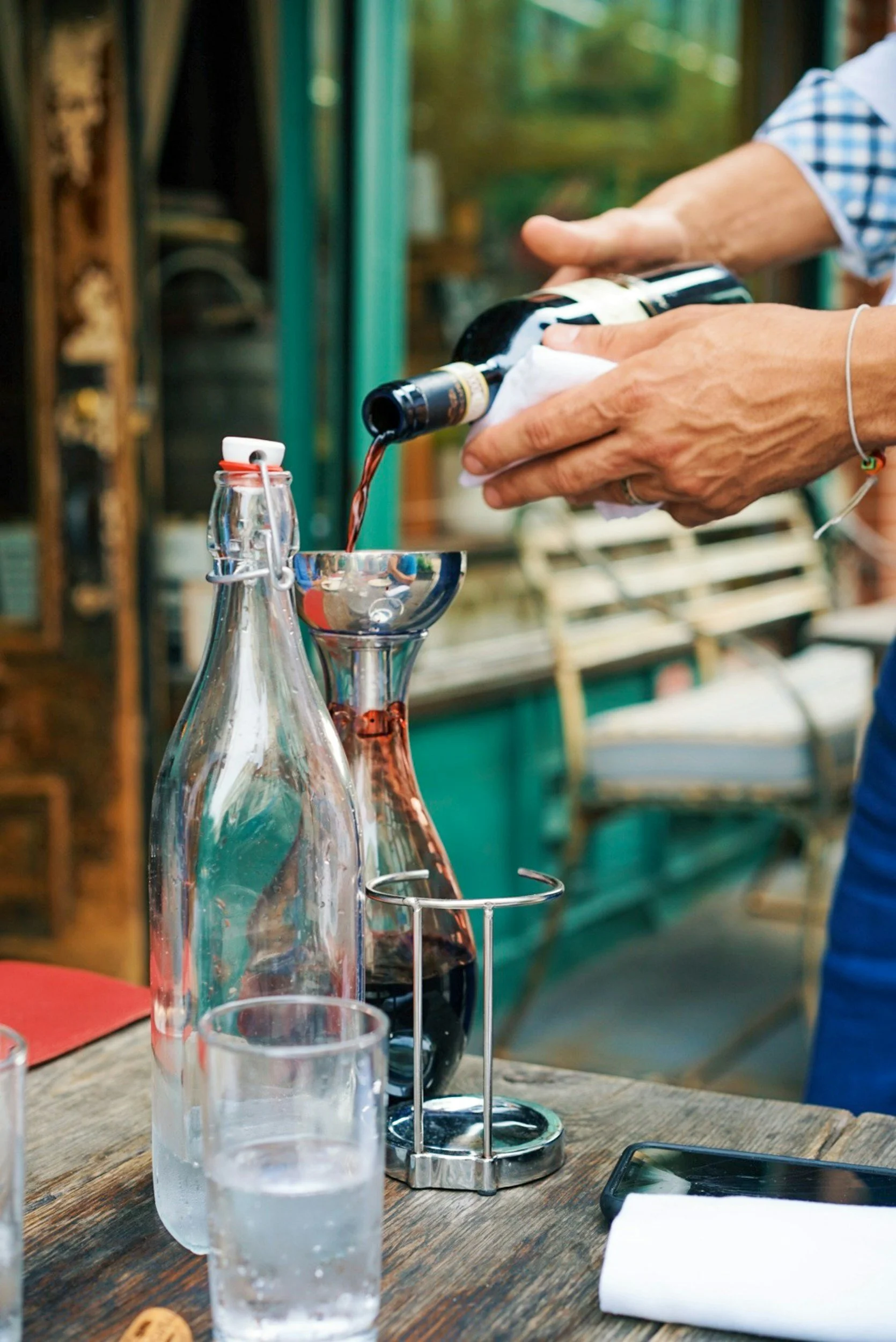 Person pouring red wine into a glass on a wooden table outside.