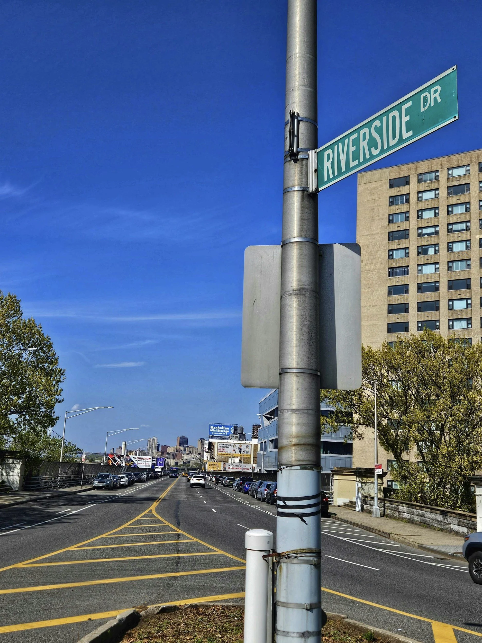 Street view with a green street sign labeled Riverside Drive attached to a metal pole, parked cars on both sides of the road, a tall beige building, trees, and billboards in the background under a blue sky.