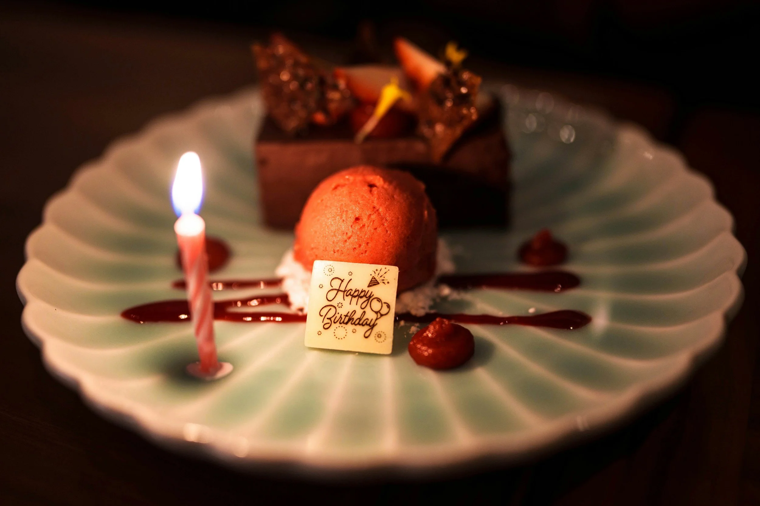 A dessert plate with a small candle, a chocolate dessert, a scoop of ice cream with a ''Happy Birthday'' sign, and decorative sauces.